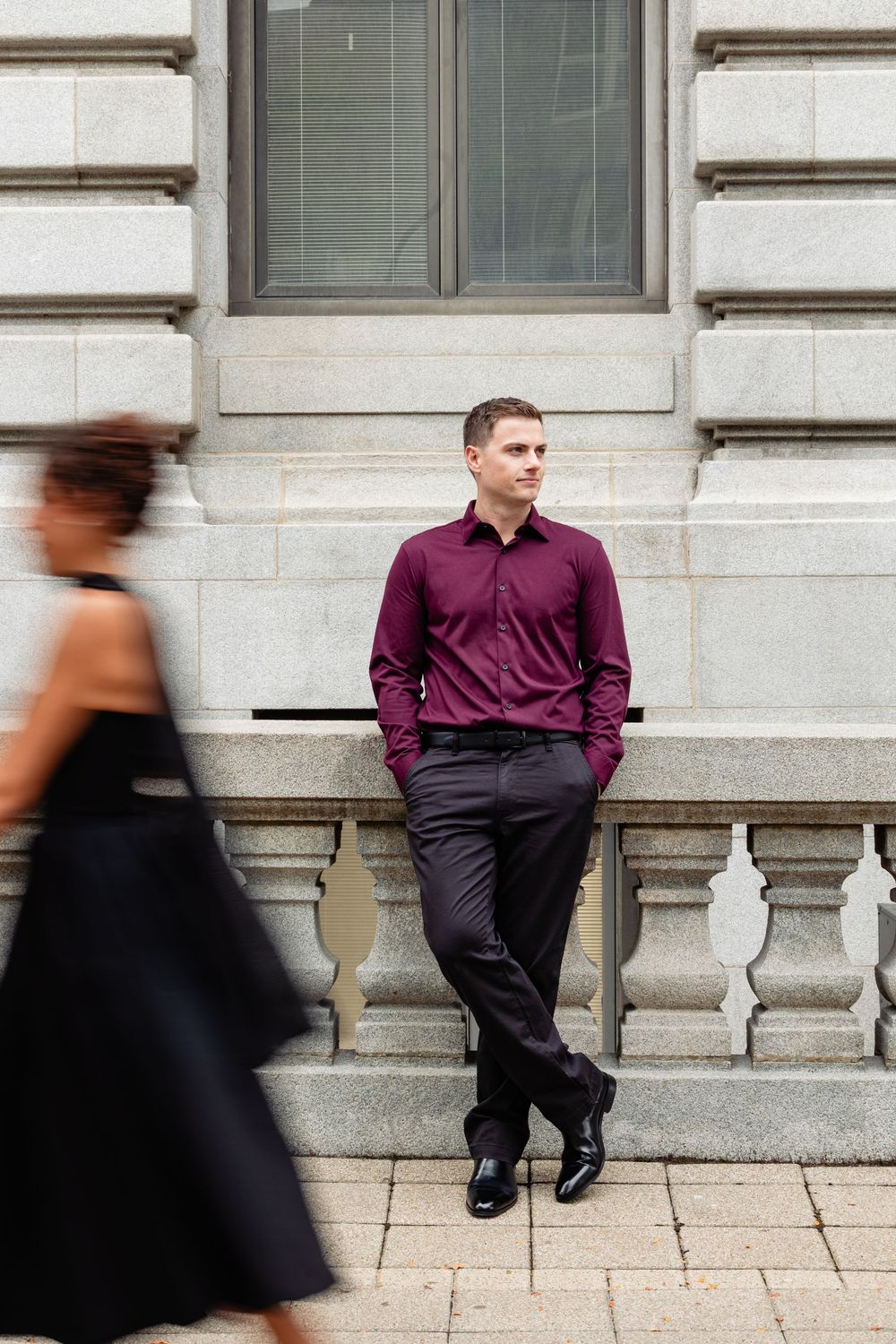 A man in a burgundy shirt and black pants leans against a stone balustrade outdoors.