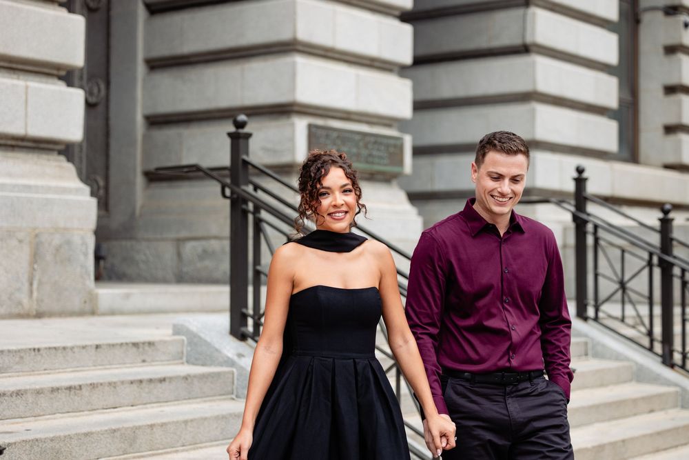 Couple in elegant attire walking down stone steps, woman in black strapless dress, man in burgundy shirt.