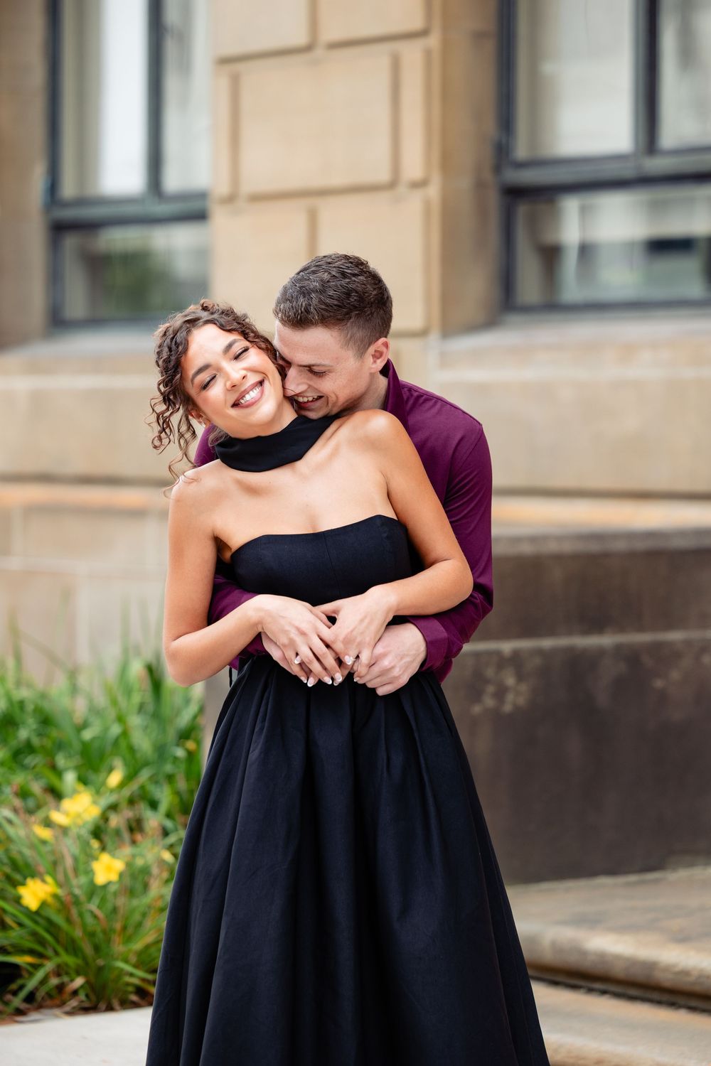 A couple embraces outdoors, woman in black strapless gown smiling joyfully as man in burgundy shirt holds her close.