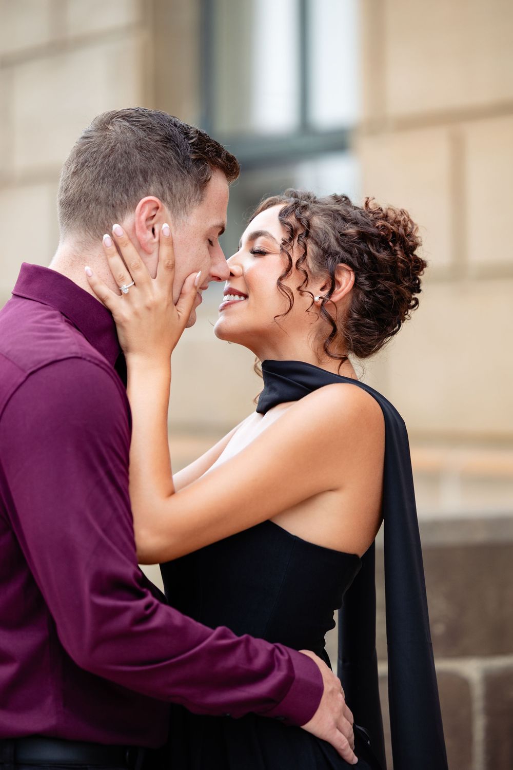 A couple shares a romantic almost-kiss moment, woman in black dress smiling, man in burgundy shirt, outdoors.