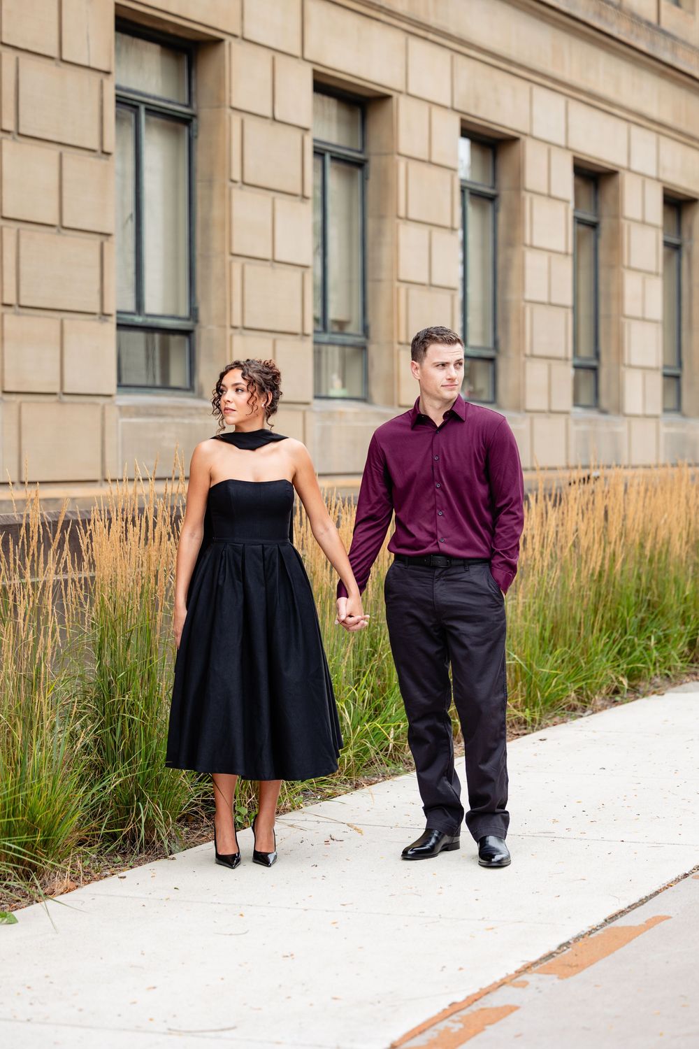 Elegant couple holding hands outdoors, woman in black strapless dress, man in burgundy shirt and black pants.