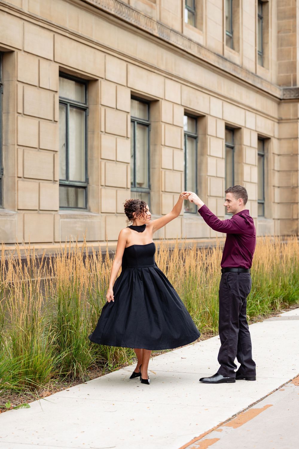 A couple dances elegantly outdoors, woman in black dress, man in burgundy shirt, beside a stone building.