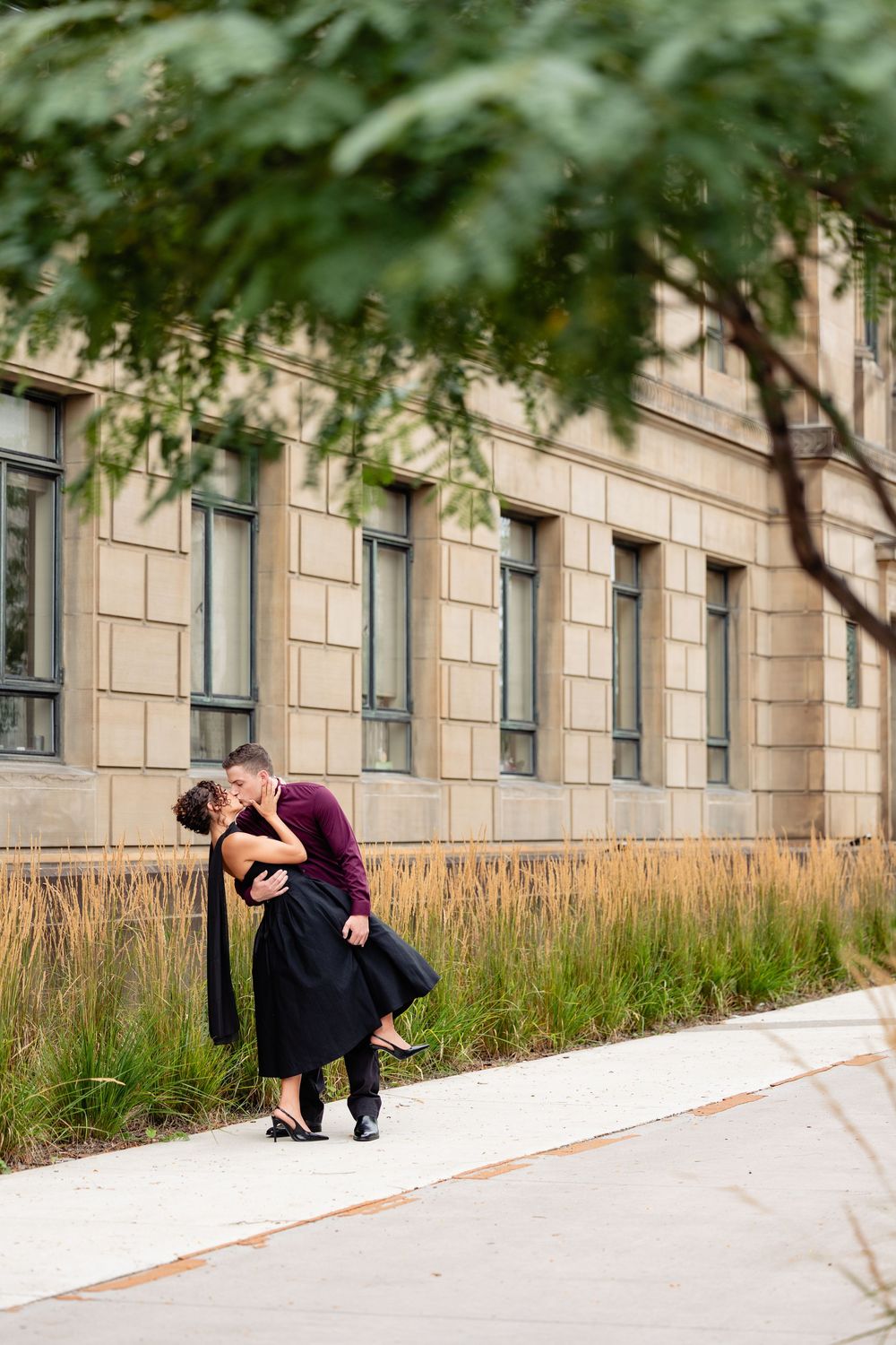 A couple shares a romantic kiss on a city sidewalk, the man lifting the woman in a black dress near a stone building.