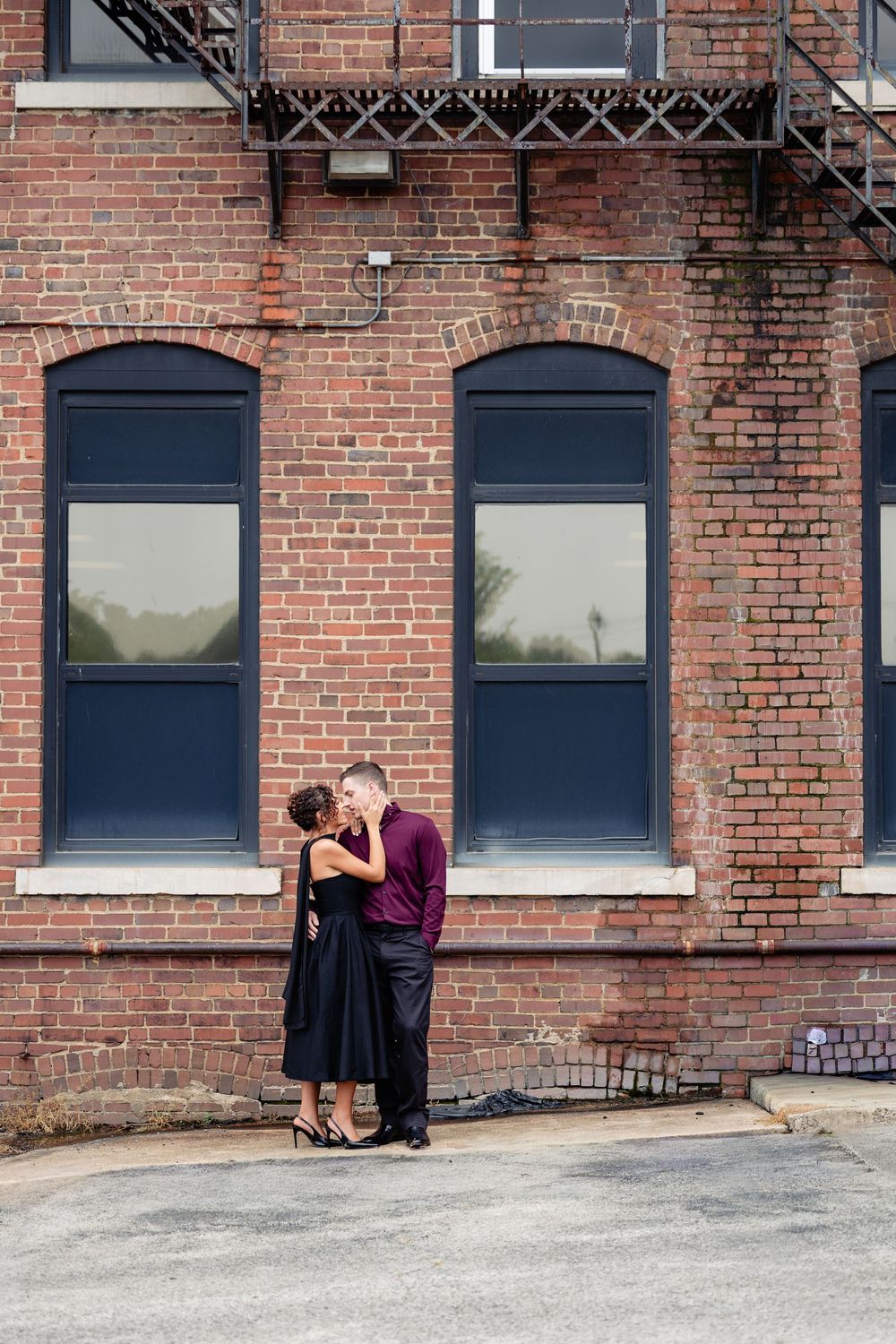 Couple sharing a romantic kiss in front of a red brick building with arched windows and fire escape.