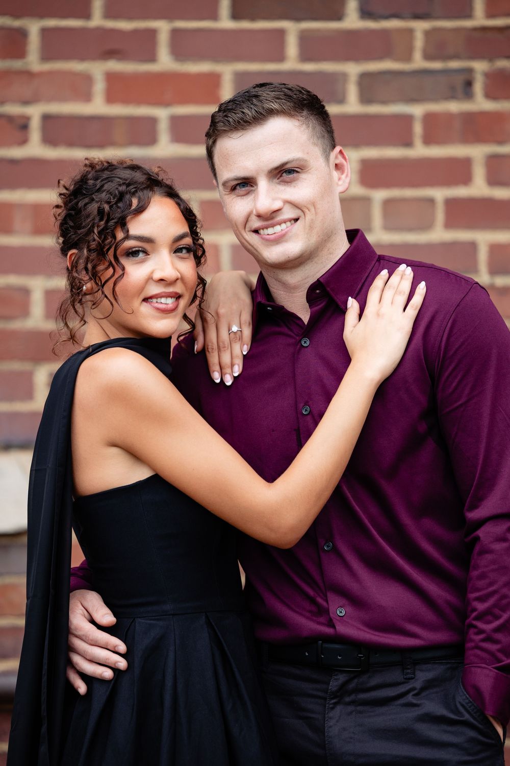Smiling engaged couple poses against brick wall, woman in black dress showing ring, man in burgundy shirt.