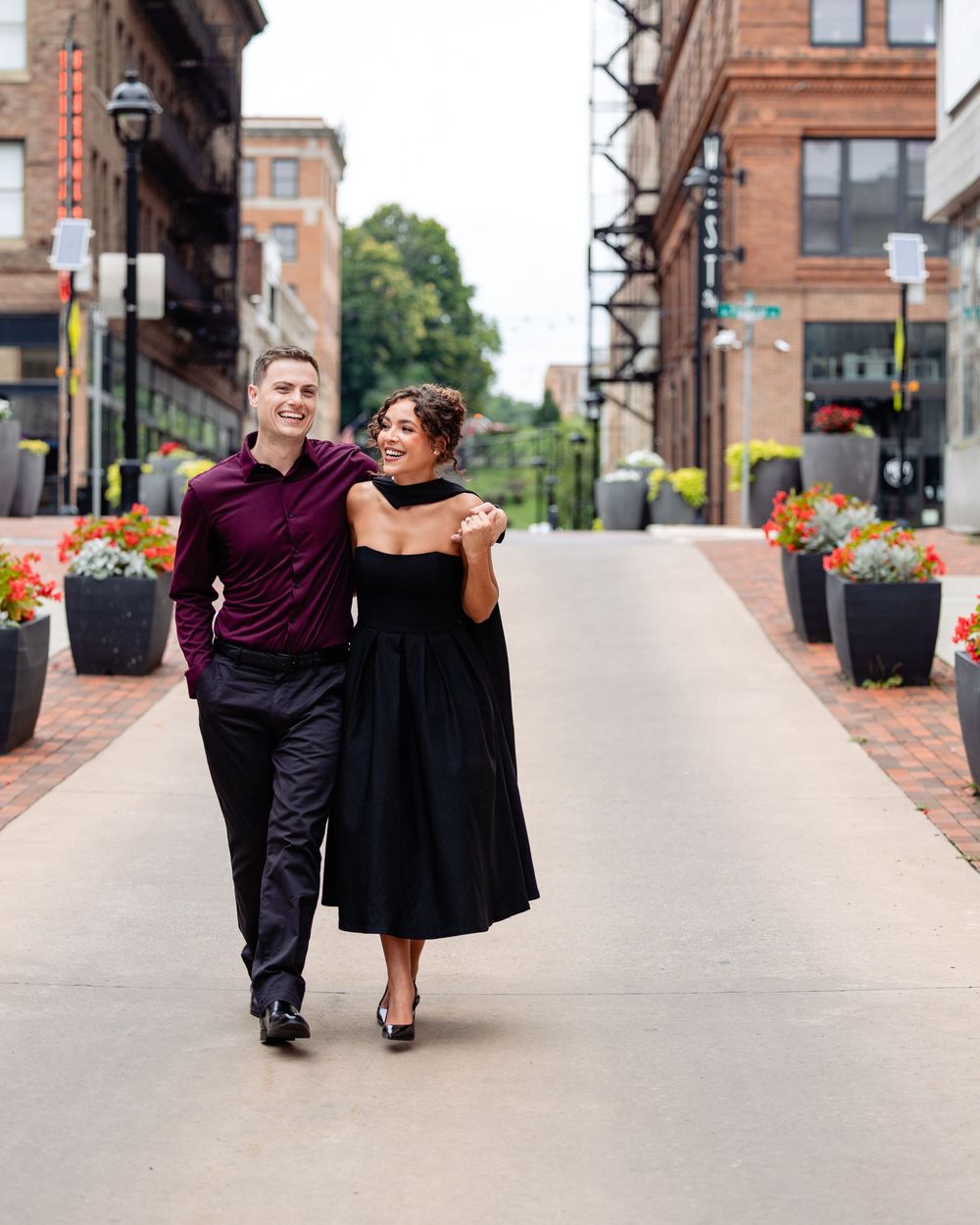 Couple walking and laughing together on a brick-lined urban street, woman in black dress, man in burgundy shirt.