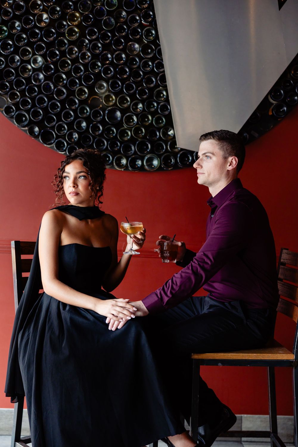 Elegant couple holding wine glasses at a romantic restaurant with wine bottle wall decor.