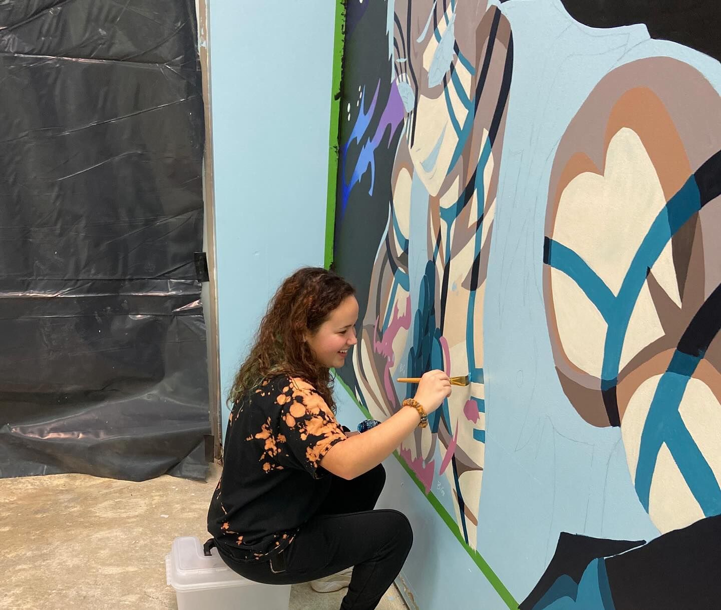 Woman with curly hair painting a colorful mural on a light blue wall, crouching with a paintbrush.