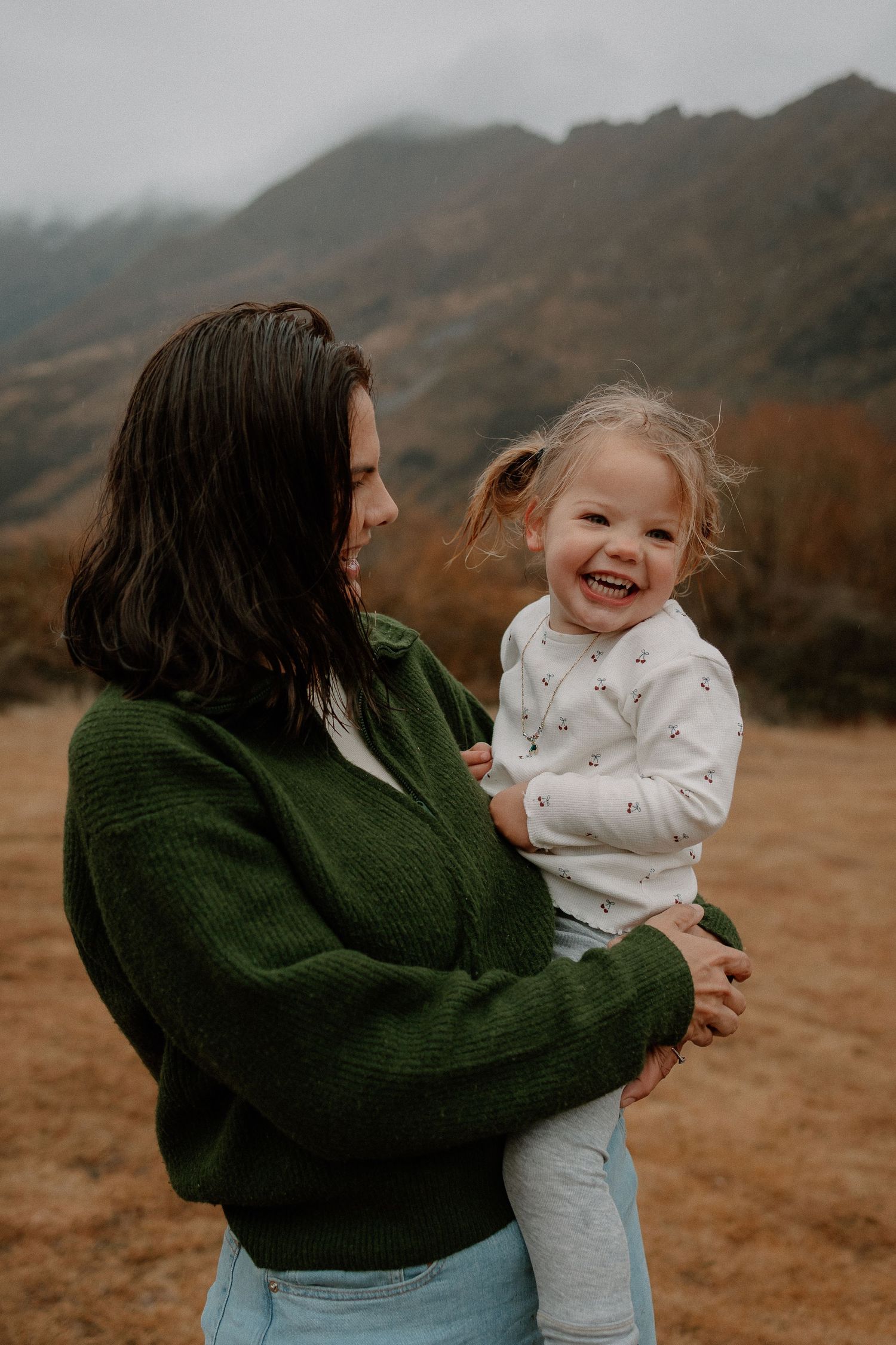 A smiling toddler girl held by a woman in a green sweater with misty mountains in the background.