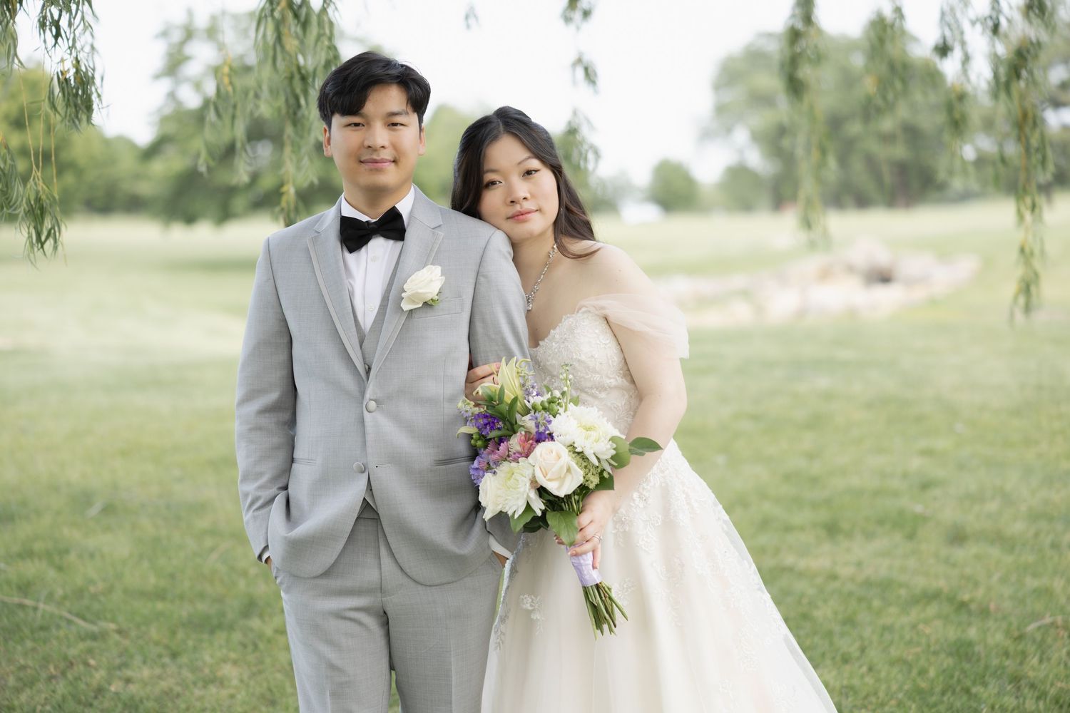 A bride and groom smile together outdoors near a weeping willow tree, bride holding a colorful bouquet.