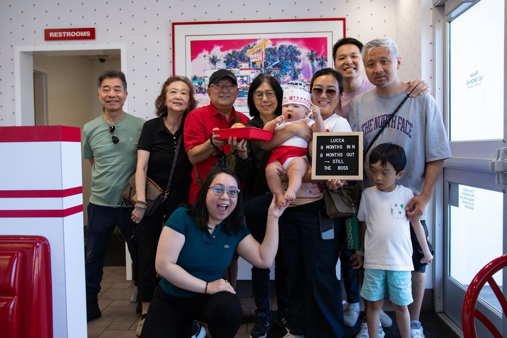 A joyful multigenerational Asian family poses together inside a diner, with one member holding a baby and a letter board.