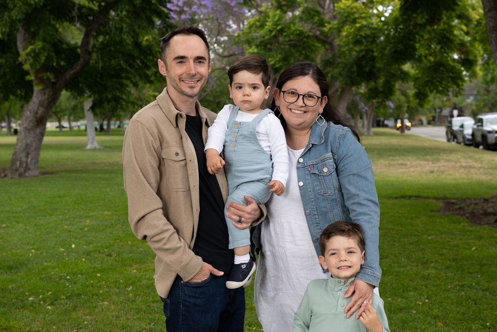 Happy family of four posing outdoors in a green park, parents holding two young children on a sunny day.