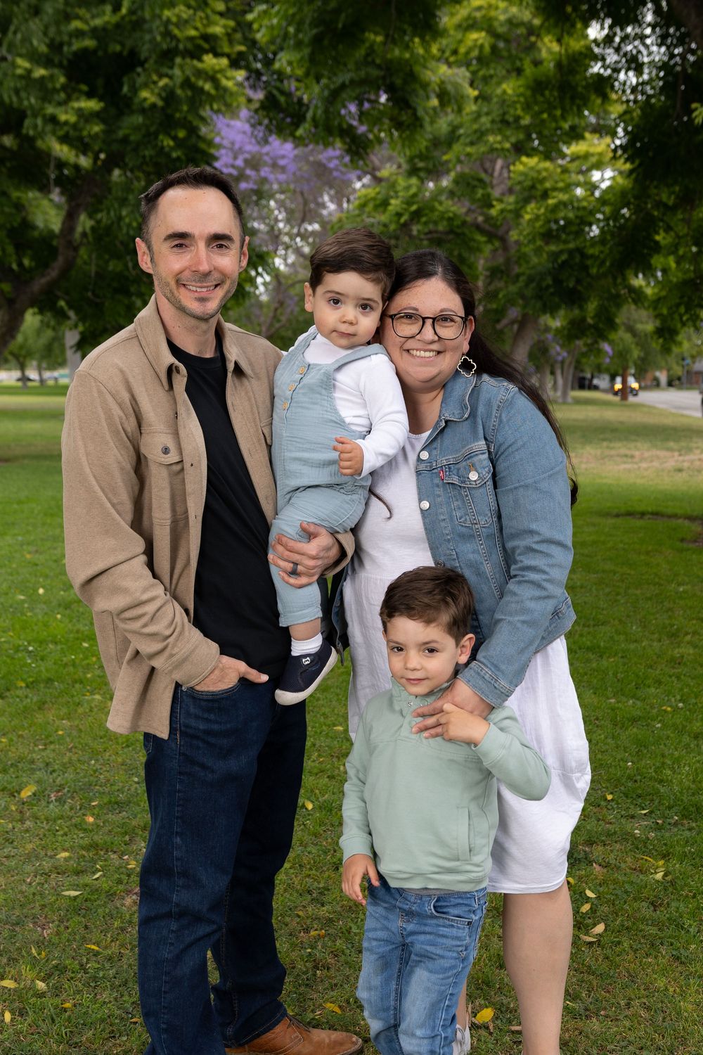 Happy family of four posing outdoors in a lush green park, parents with two young boys smiling for a portrait.