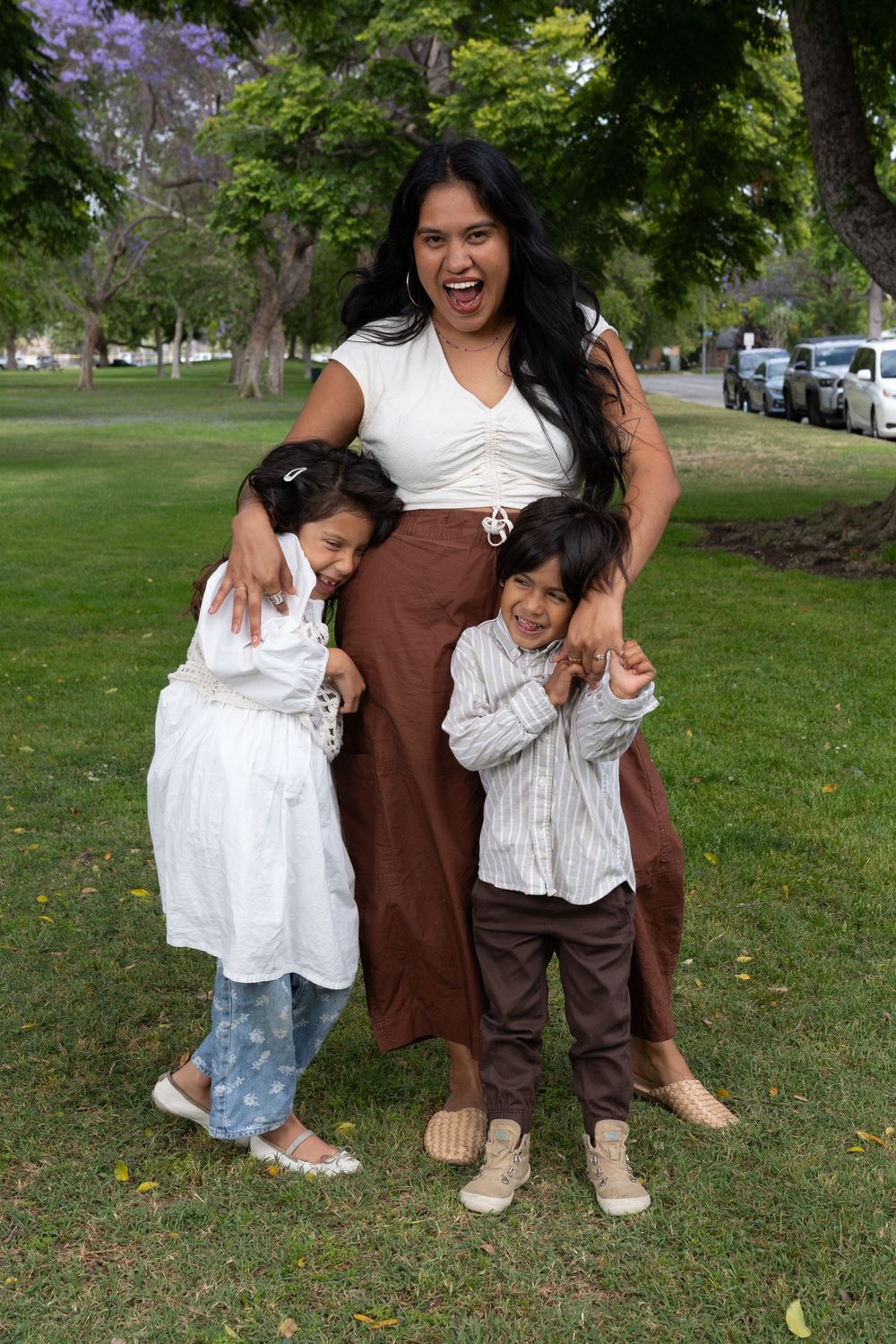 A joyful woman in white top and brown skirt hugs two young children while posing in a sunny green park.