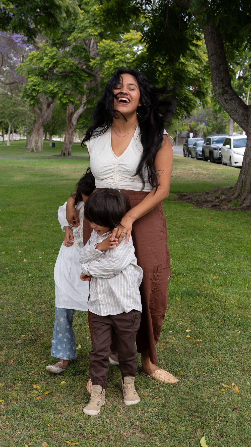 A laughing woman in white top and brown skirt hugs two young children on green park grass with trees behind.