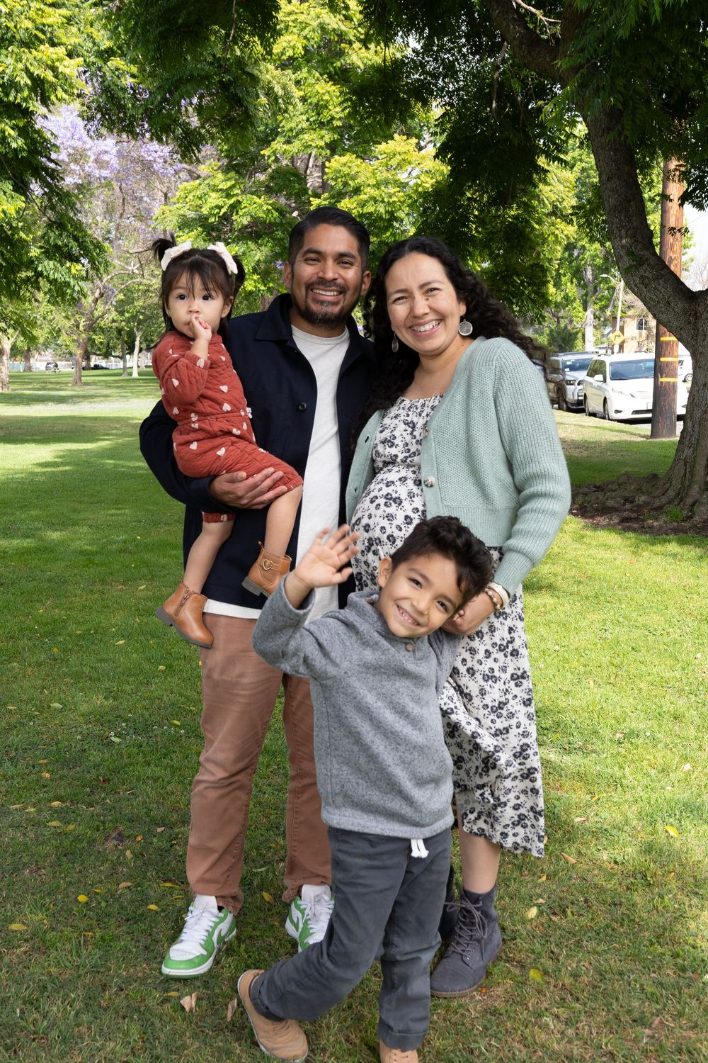 Happy family of four posing outdoors in a green park, parents smiling with two young children on a sunny day.