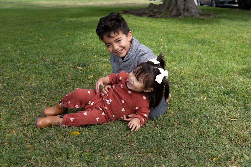 Two young children playing on green grass in a park, boy in gray smiling at toddler girl in red floral outfit.