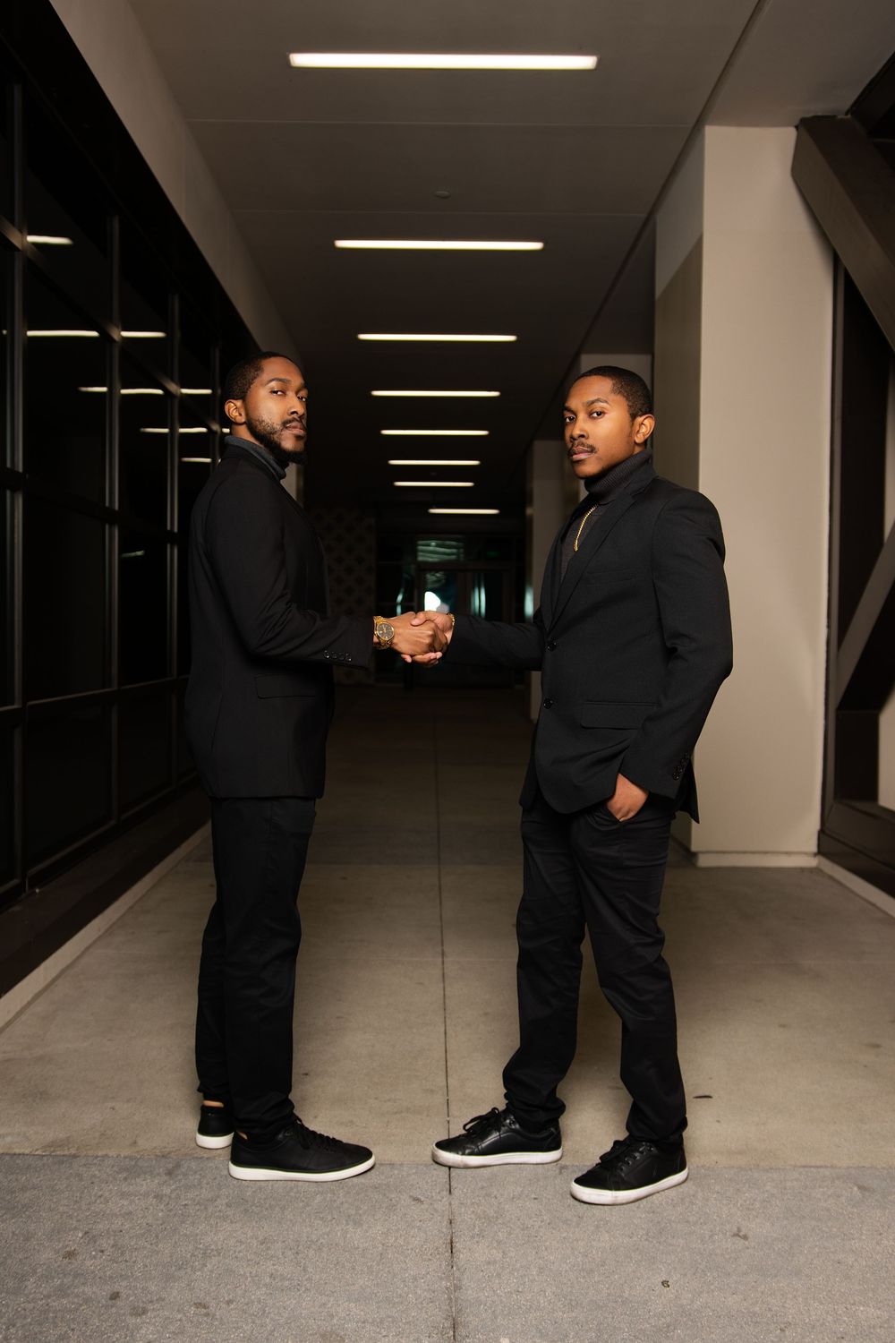 Two young men in black suits shaking hands in a modern hallway corridor with overhead lighting.