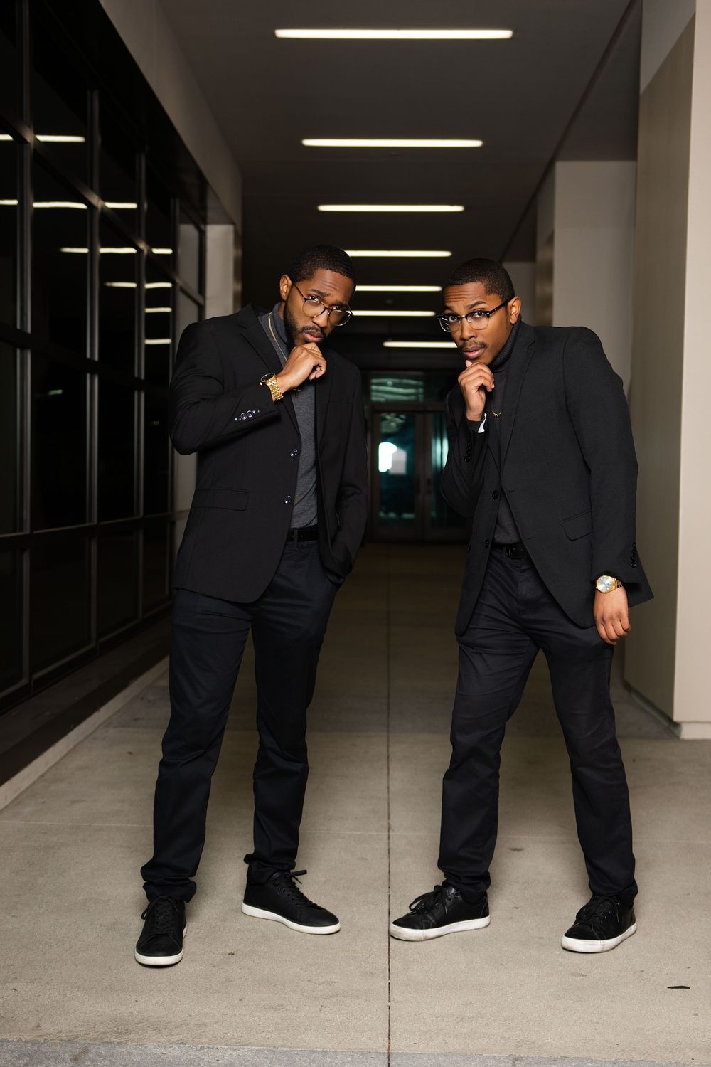 Two stylish men in black suits pose confidently in a modern hallway with ceiling lights.