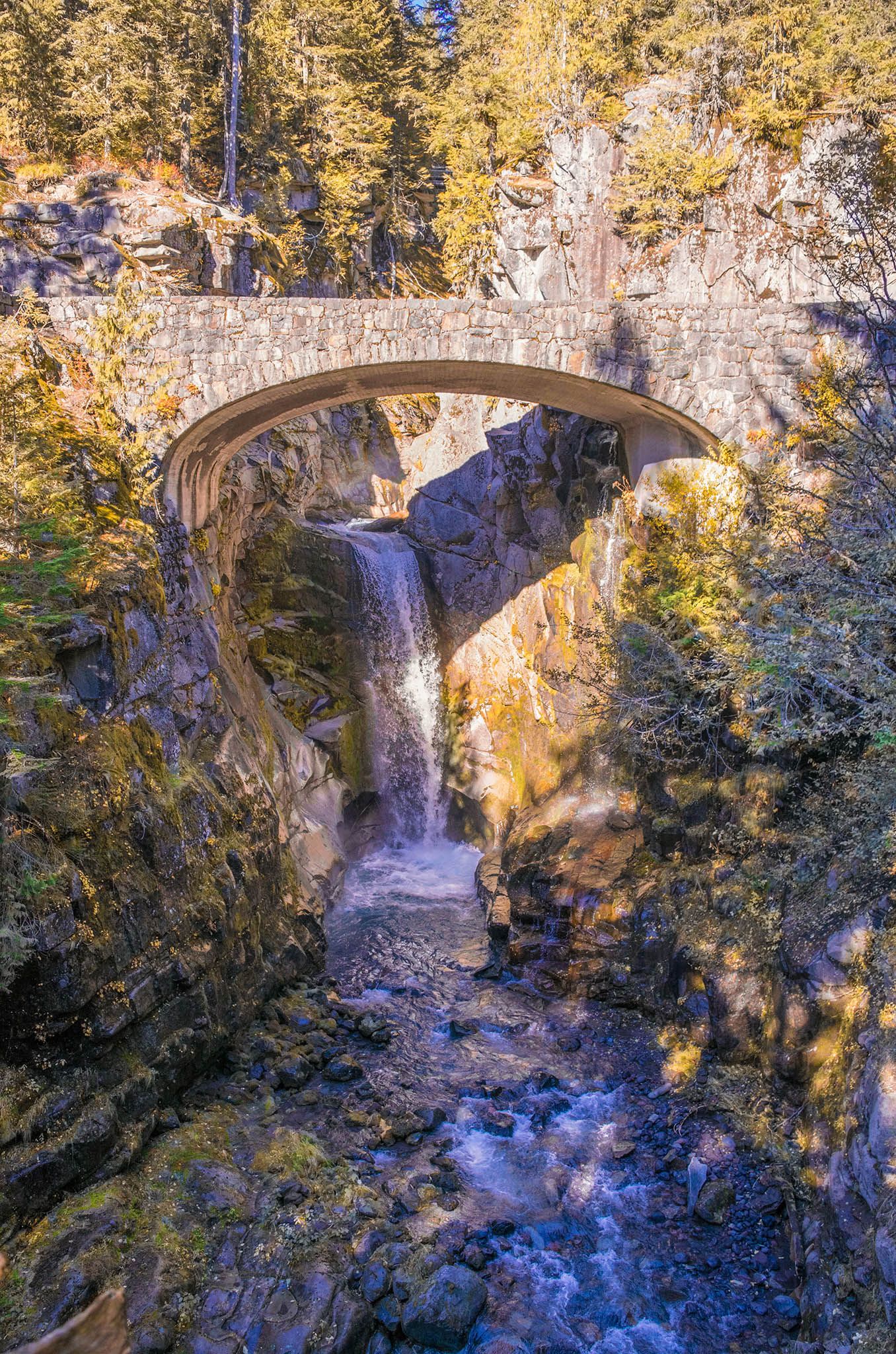Historic stone arch bridge spans across dramatic waterfall in autumn forest setting.