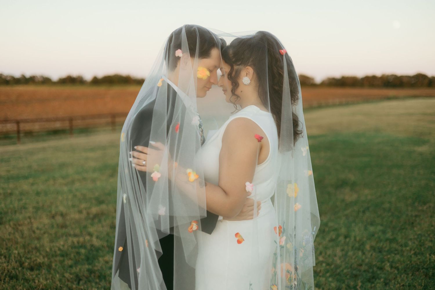 Bride in white sleeveless gown with tattoo stands before groom in navy suit at golden hour farm backdrop.