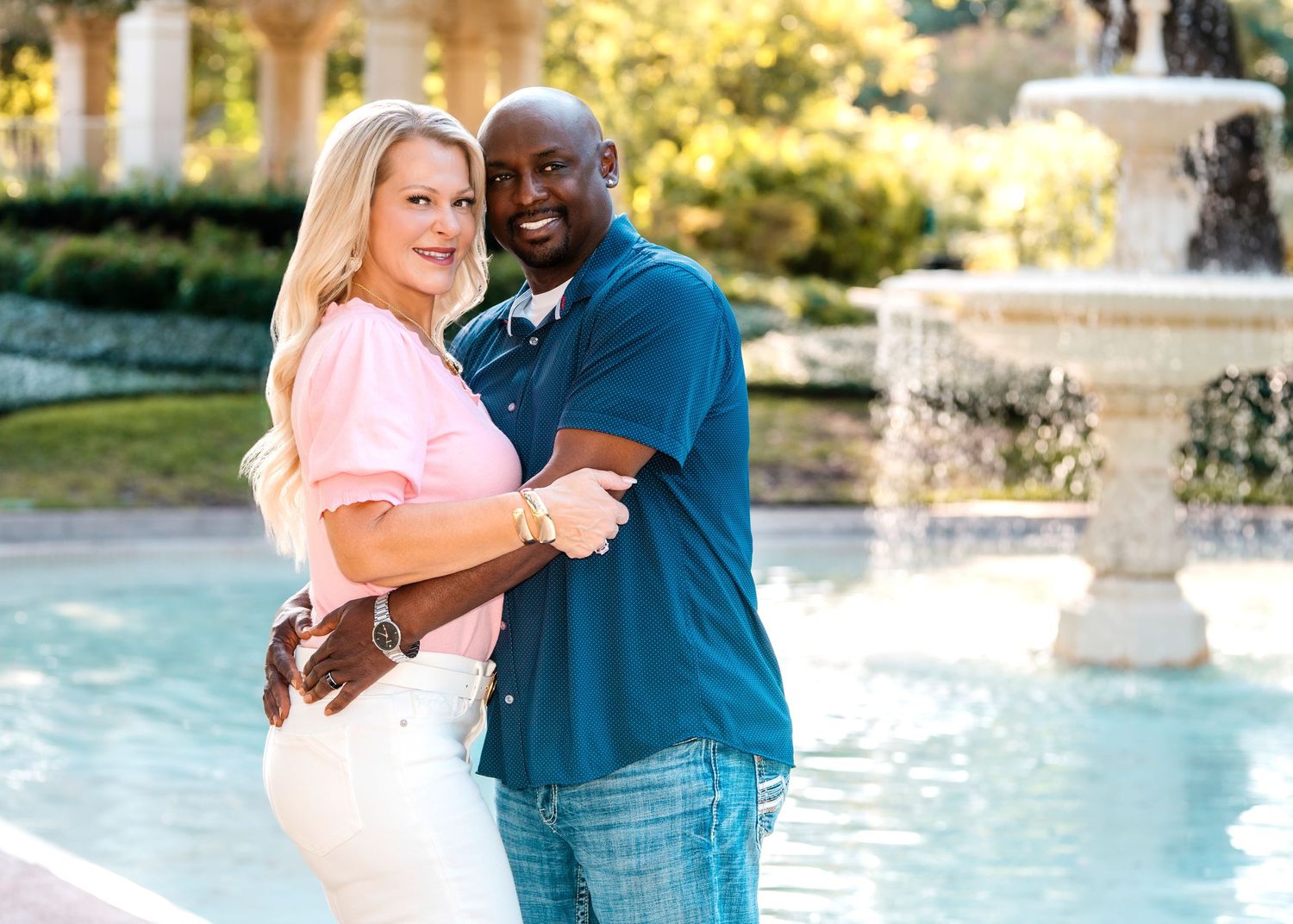 A smiling couple embraces near an elegant fountain outdoors, woman in pink top and man in blue shirt.