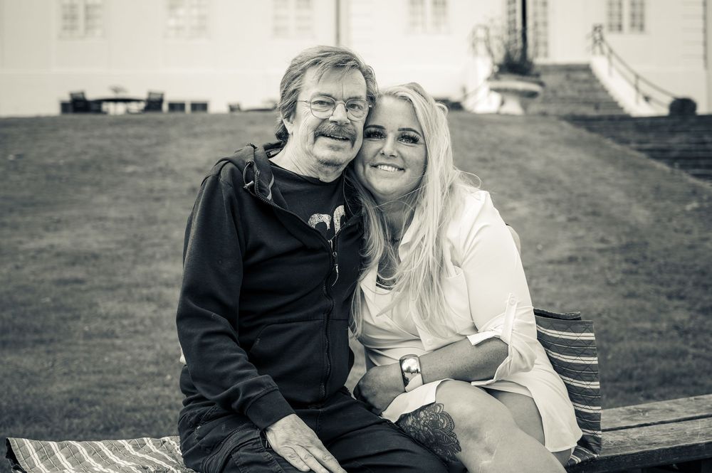 Black and white photo of a smiling couple sitting together on a bench outdoors near a white building.