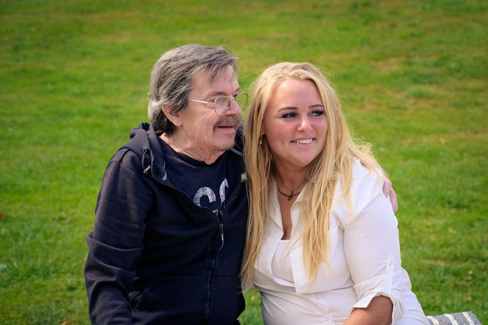 An older man with a mustache and a young blonde woman smiling together outdoors on green grass.