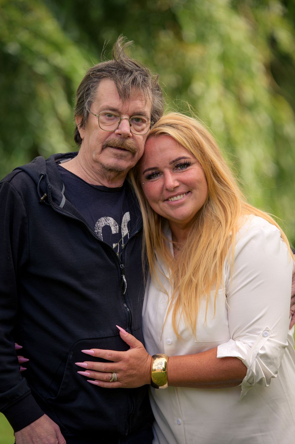 An older man with glasses and mustache poses with a smiling blonde woman in a white blouse outdoors.