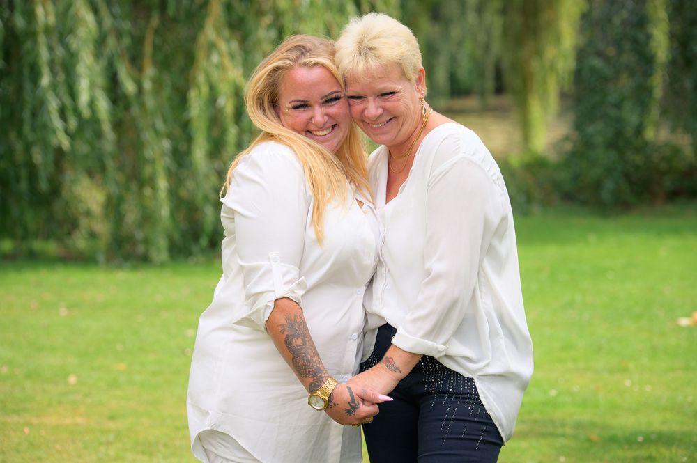 Two women in white blouses smiling and embracing outdoors with green willow trees in the background.
