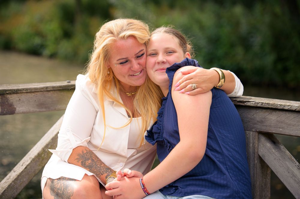 Two women embracing warmly on a wooden bridge outdoors, sharing a tender moment near a pond.