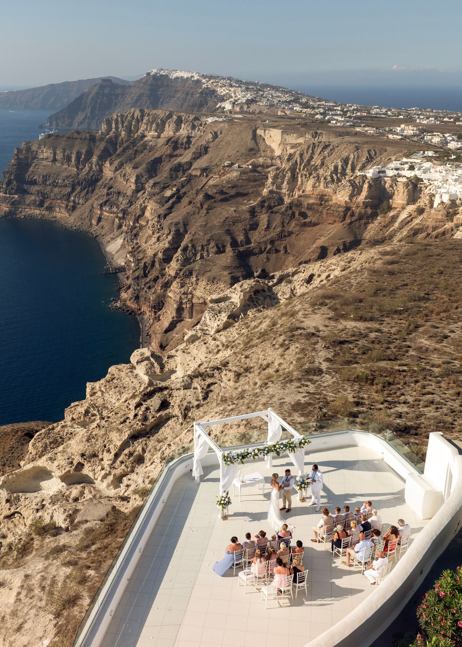 Outdoor wedding ceremony on a cliffside terrace in Santorini, Greece, with a traditional windmill and stunning Aegean Sea views.