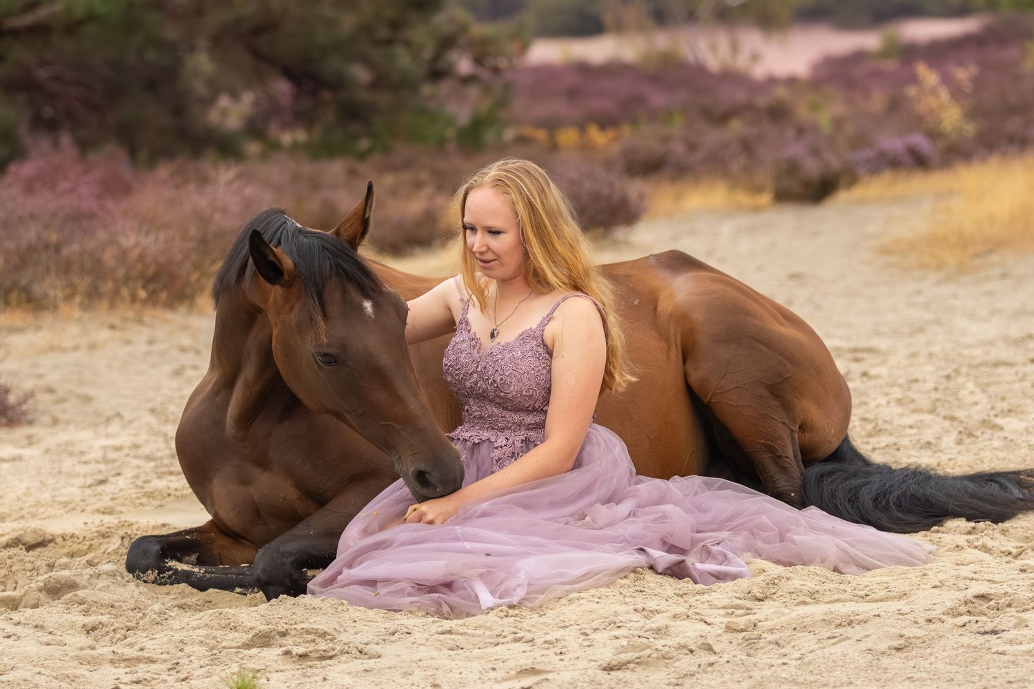 Vrouw in een lange roze jurk zit naast een liggend bruin paard op een zandvlakte bij de heide.