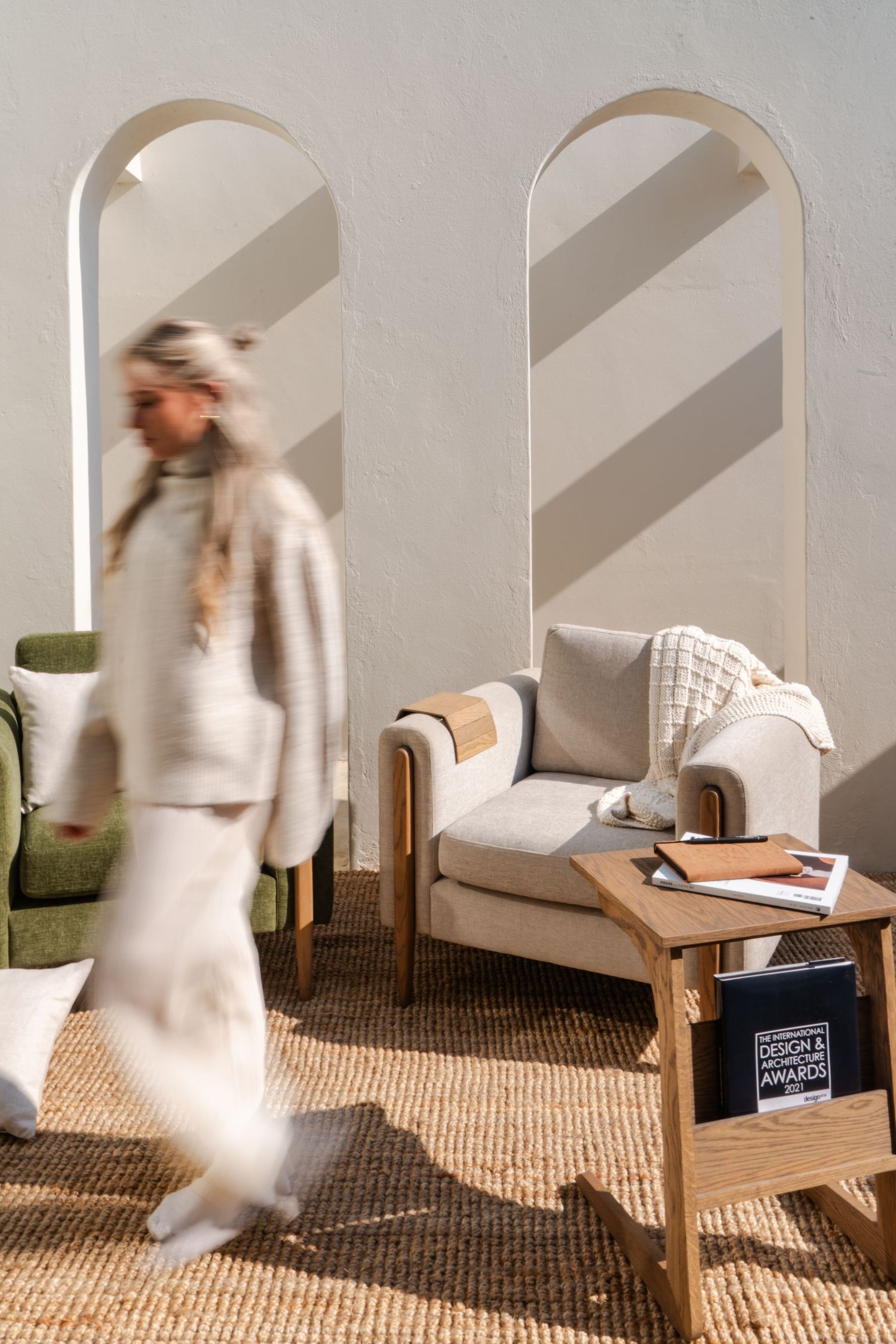 Blurred woman in cream outfit walks past arched walls and neutral armchair in minimalist scene during a lifestyle product shoot.