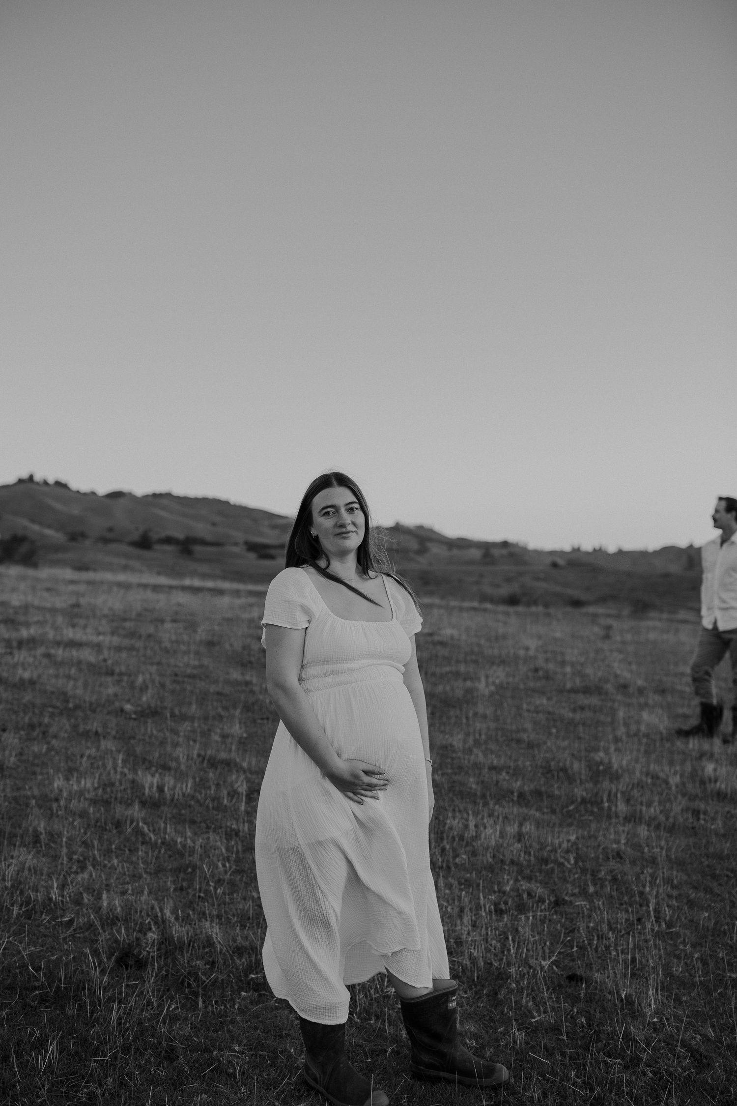 Black and white maternity photo session on farm. Tasman. Mum to be holding bump in white dress and gumboots.