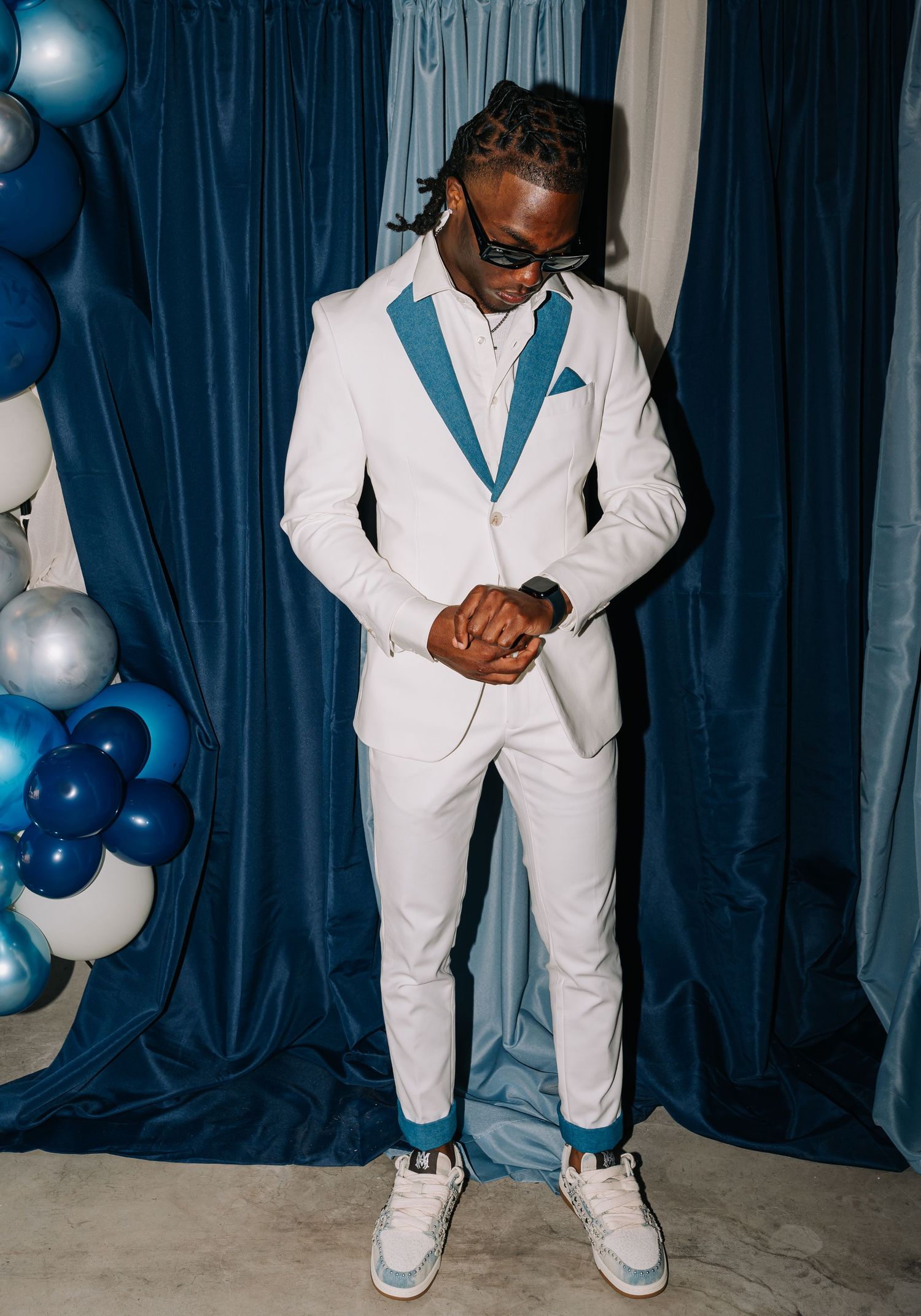Man in white suit with blue lapels and pocket square, wearing sunglasses and sneakers, posing near blue balloons.