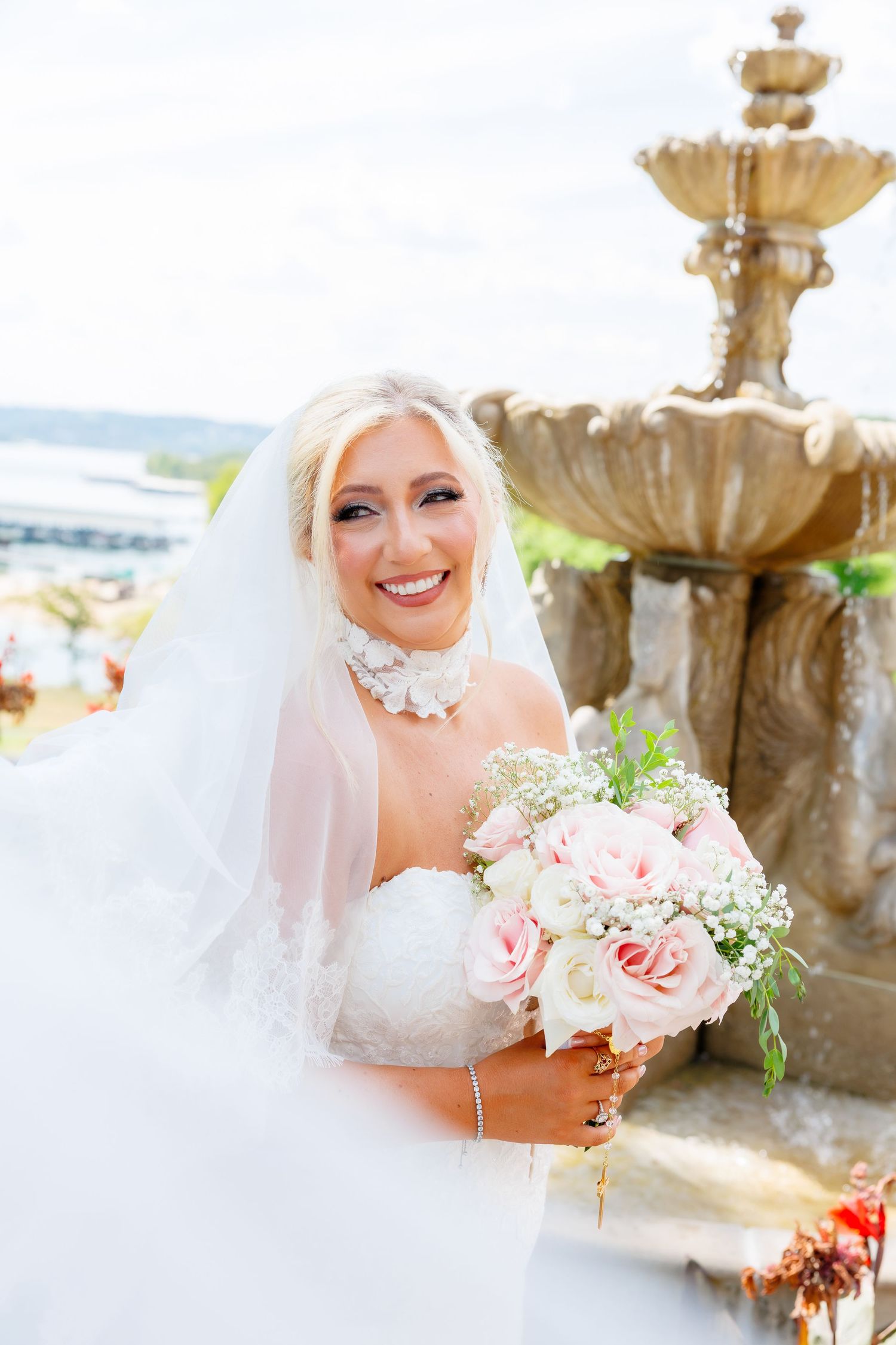 Bride in white gown and veil holds bouquet under flowering arch in lush garden setting.