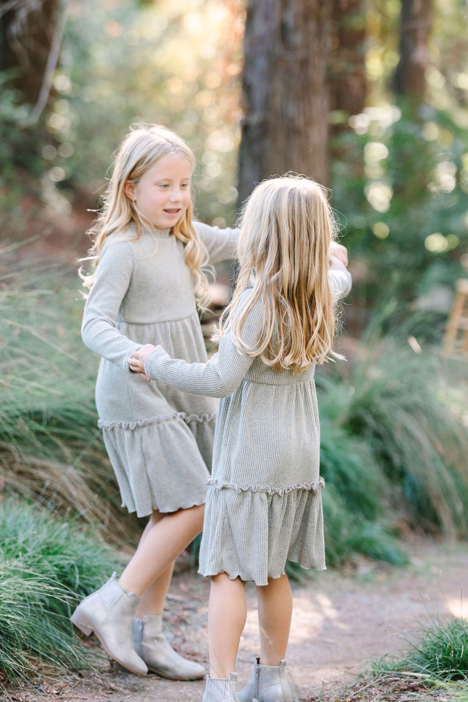 Two young girls in matching gray dresses and boots dancing together on a forest path at uc Davis arboretum mini session with Jessica Heuer