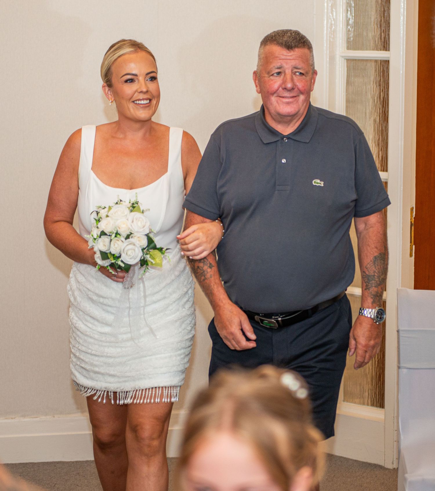 Bride in white mini dress holding bouquet walks with older man in gray polo shirt at wedding ceremony.