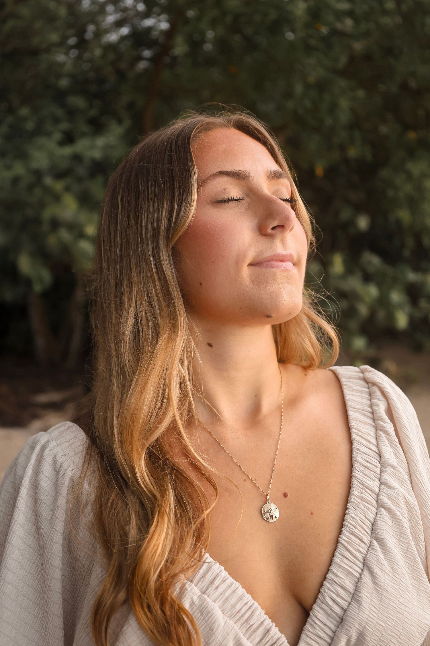 Young blonde woman with wavy hair and a pendant necklace enjoying golden hour sunlight with eyes closed outdoors.