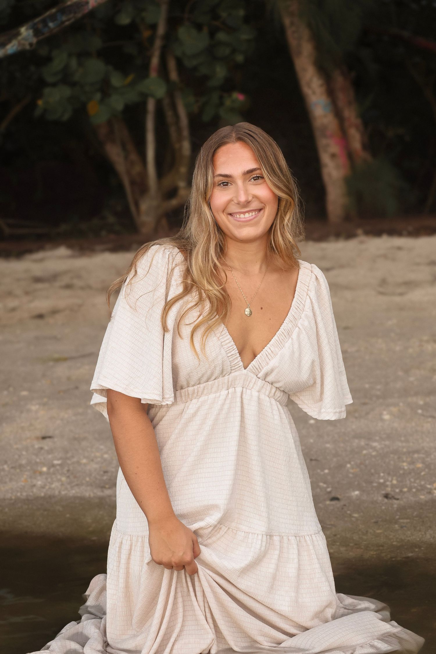 Smiling blonde woman in a white V-neck tiered maxi dress posing on a tropical beach at sunset.