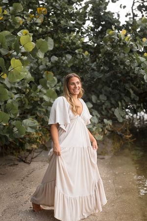 Smiling blonde woman in a flowing white boho maxi dress posing under lush green tropical foliage.