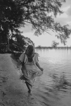 Black and white photo of a woman in a maxi dress running barefoot along a tranquil shoreline.