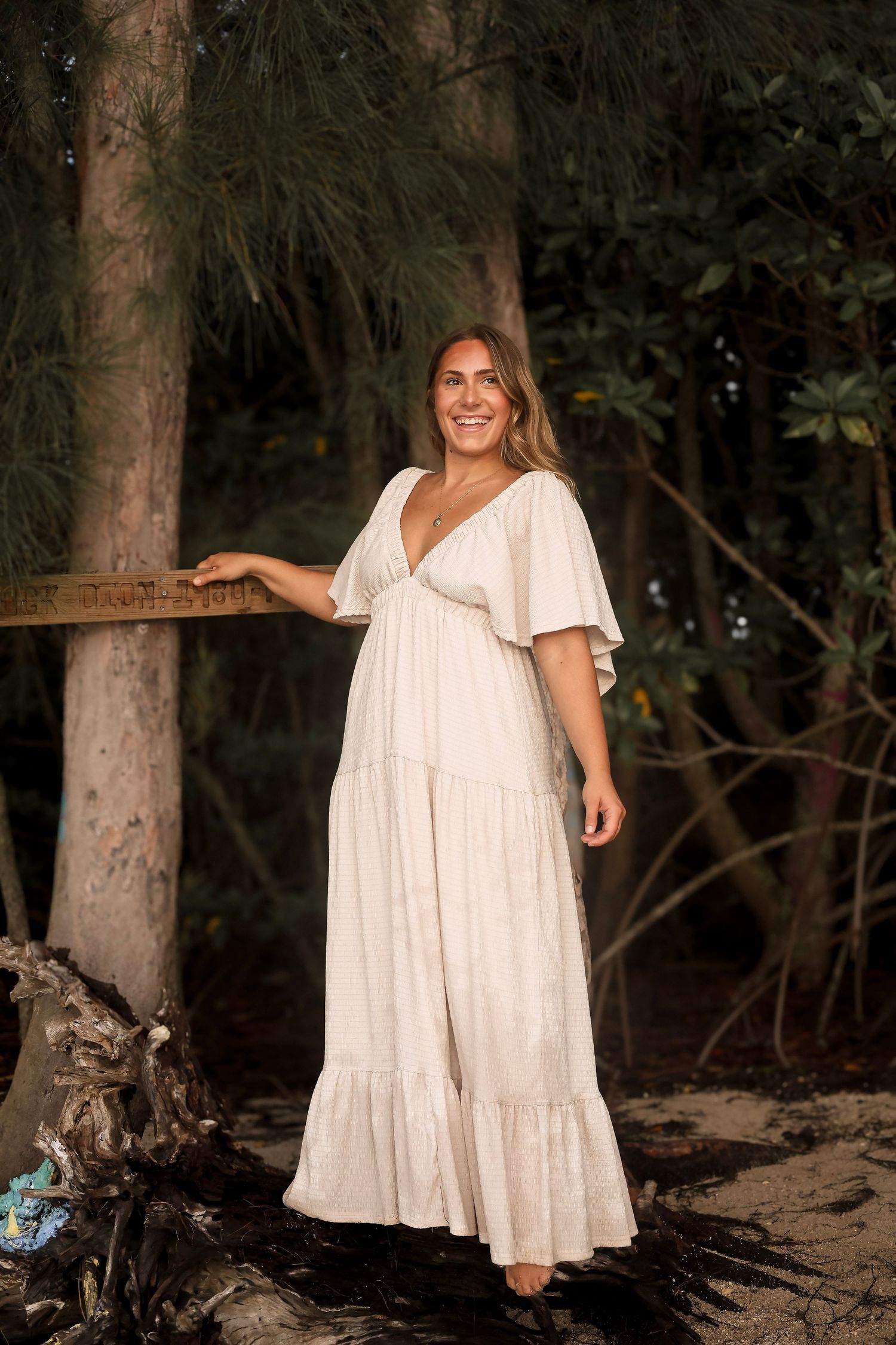 Smiling blonde woman in a flowing white boho maxi dress standing among trees on a sandy beach.