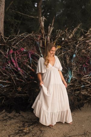 Blonde woman in a white tiered maxi dress posing near colorful decorated driftwood in a forest setting.
