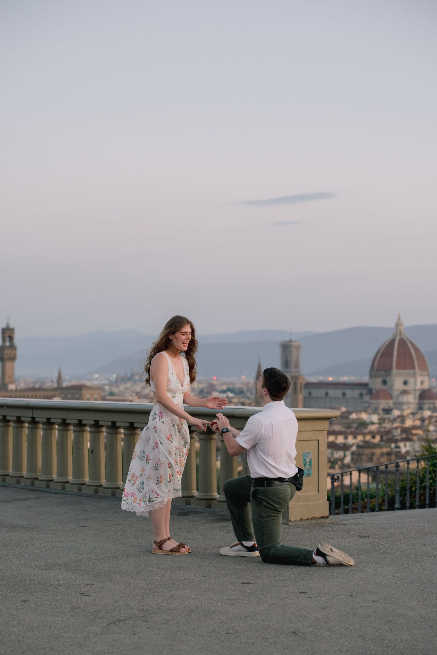 Man proposes on one knee to woman in floral dress at Piazzale Michelangelo with Florence skyline backdrop.