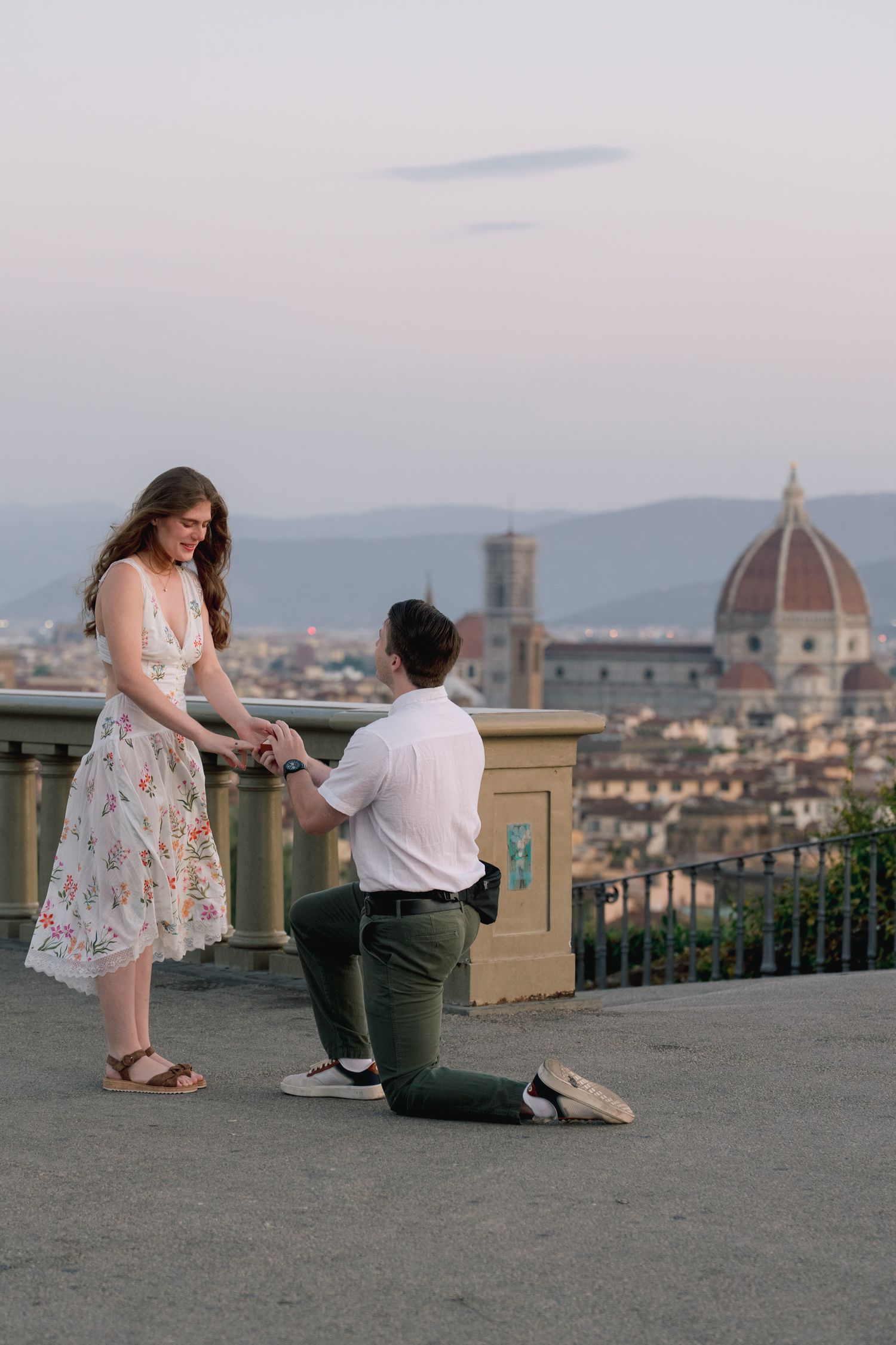 Man proposes on one knee to woman in floral dress with Florence cathedral skyline at dusk.