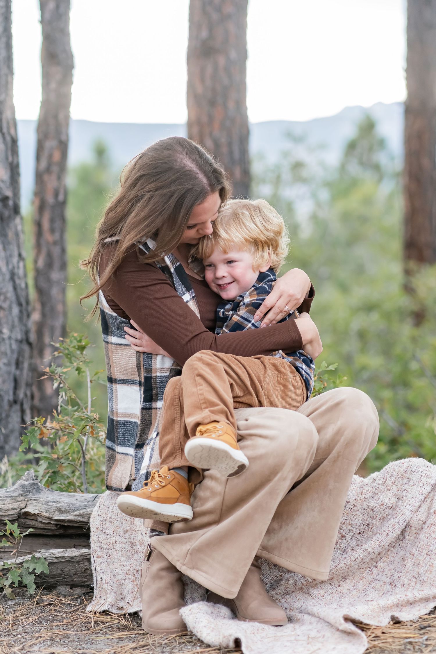 A mother hugs her laughing toddler son while sitting on a log in a forest setting during fall.