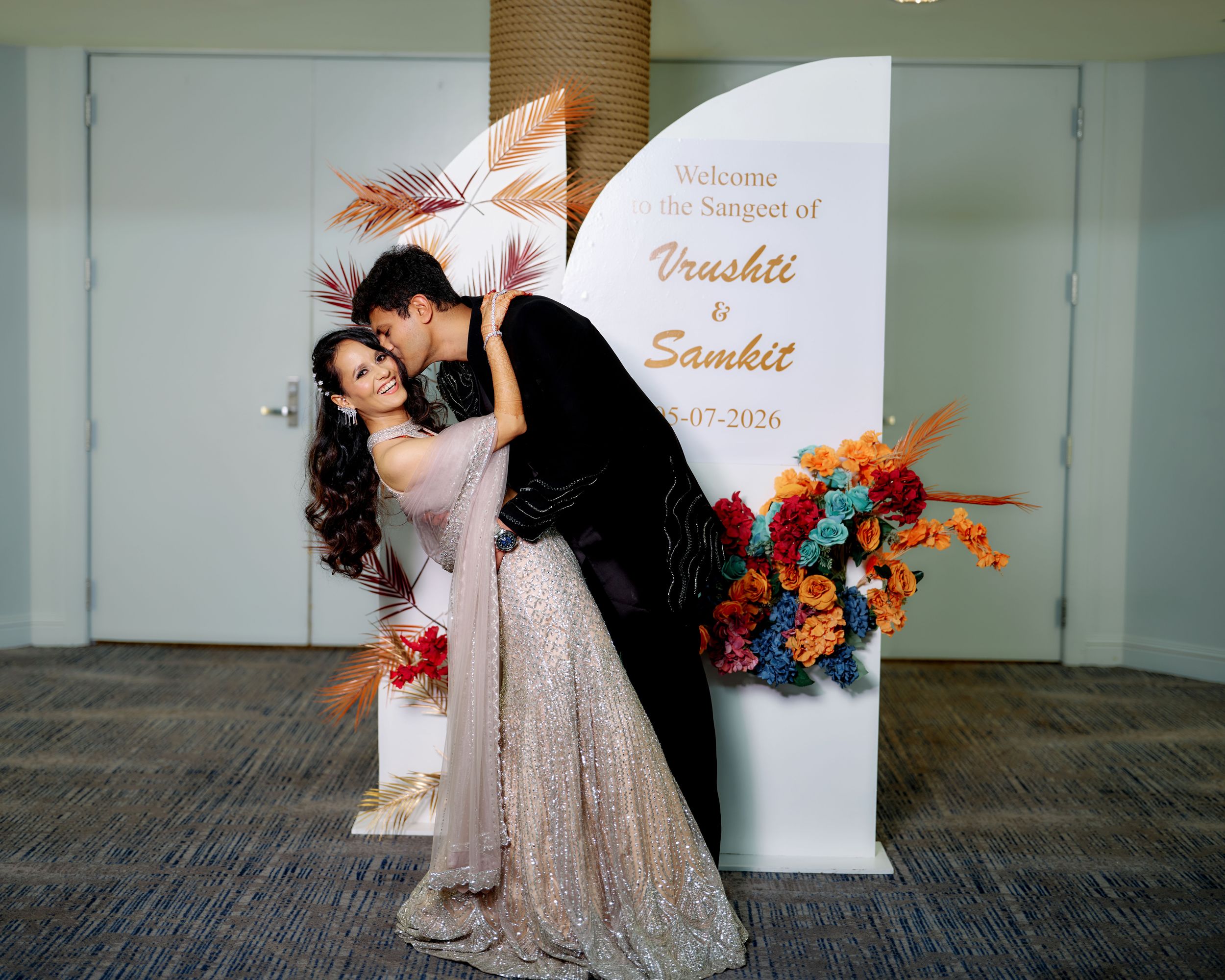 Couple posing at Vrushit and Samkit Sangeet ceremony backdrop with colorful floral decorations.