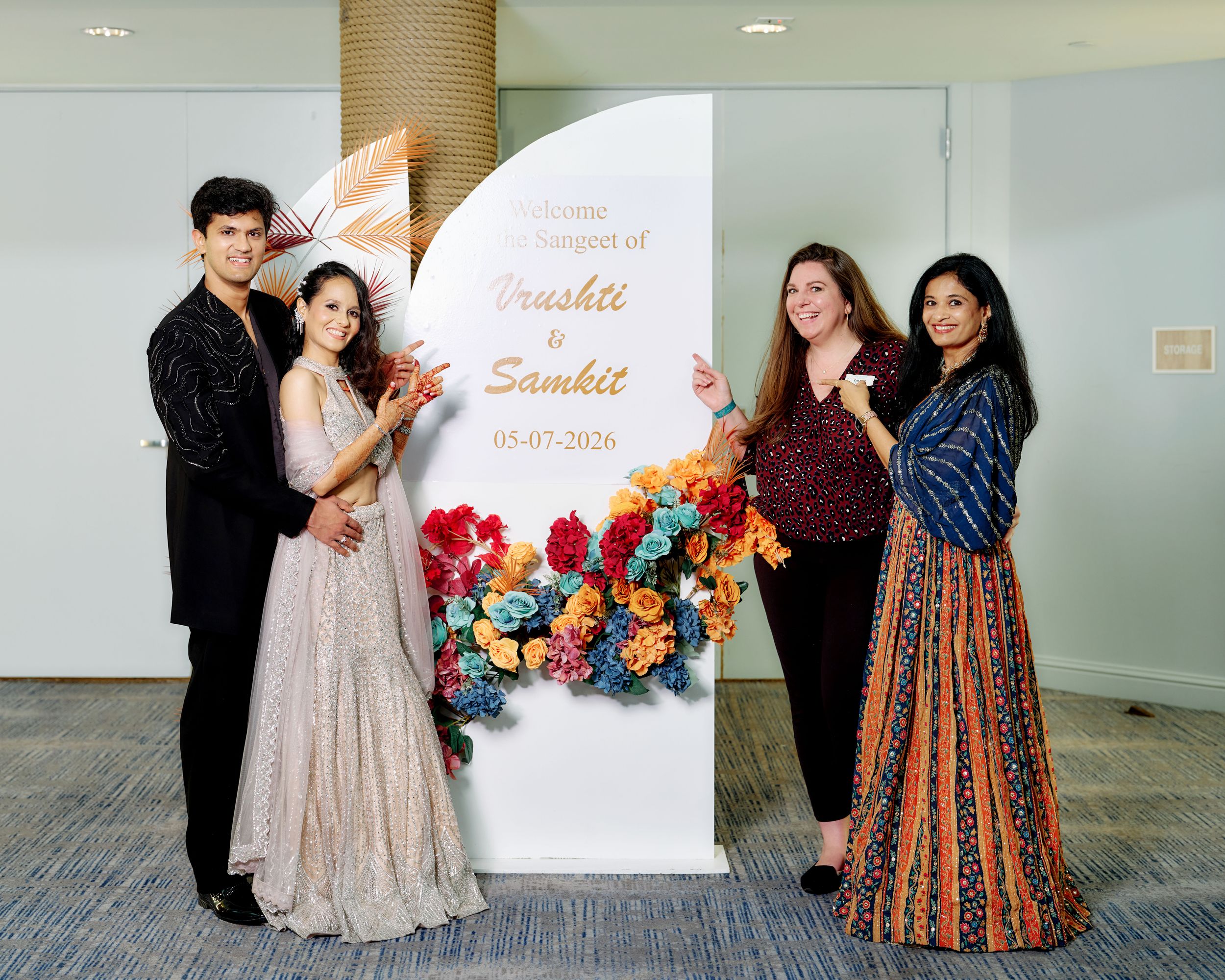 Four guests pose beside a floral wedding welcome sign for Drushti and Samhit at an elegant engagement banquet.